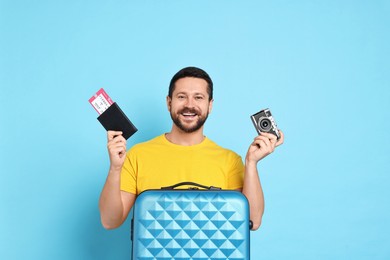 Photo of Happy man with suitcase, passport, ticket and vintage camera on light blue background