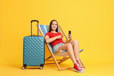 Photo of Happy young woman pointing at smartphone while sitting on folding chair and suitcase against orange background