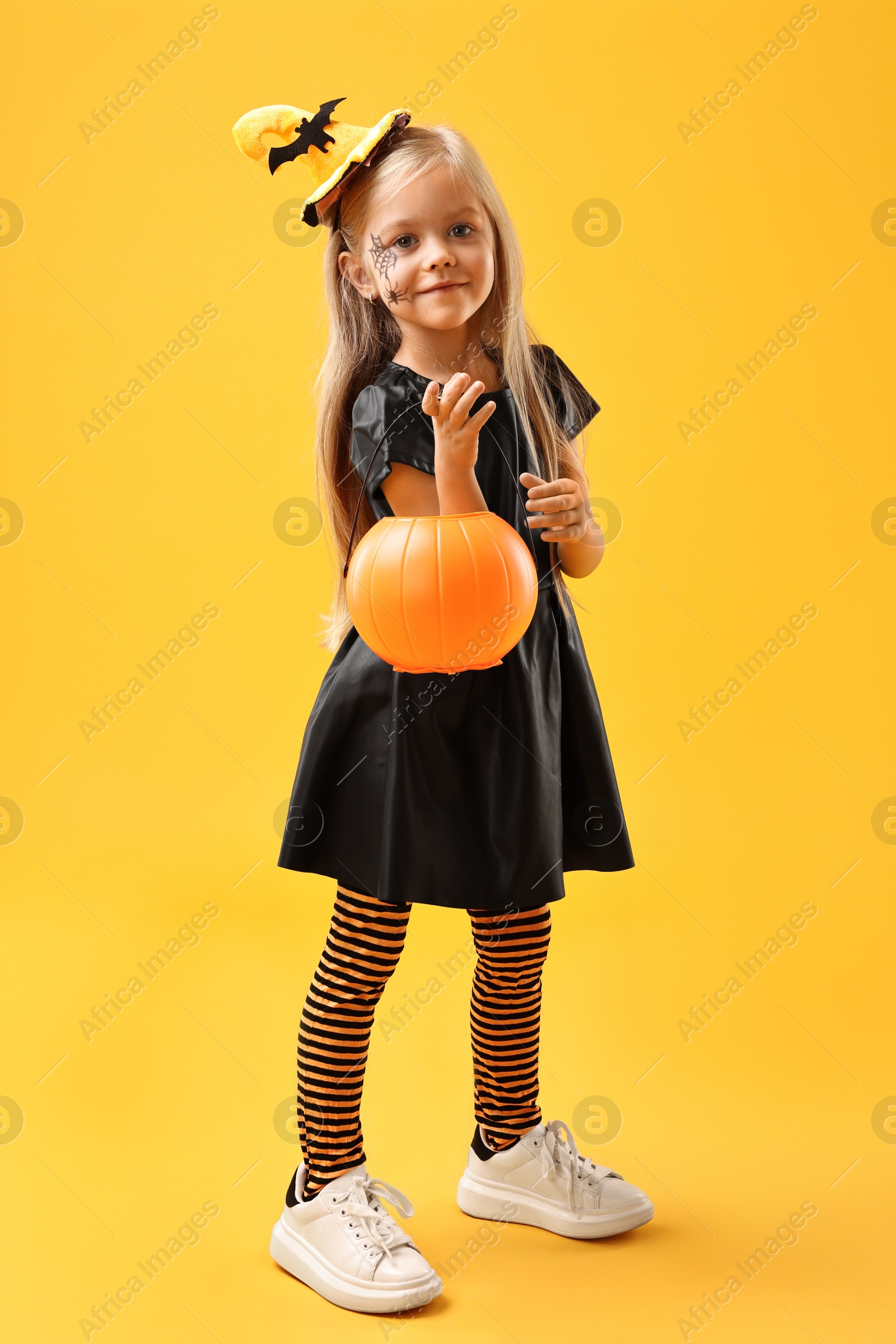 Photo of Cute girl with pumpkin bucket dressed like witch on yellow background. Halloween celebration