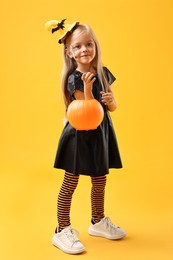 Photo of Cute girl with pumpkin bucket dressed like witch on yellow background. Halloween celebration