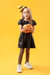 Photo of Cute girl with pumpkin bucket dressed like witch on yellow background. Halloween celebration