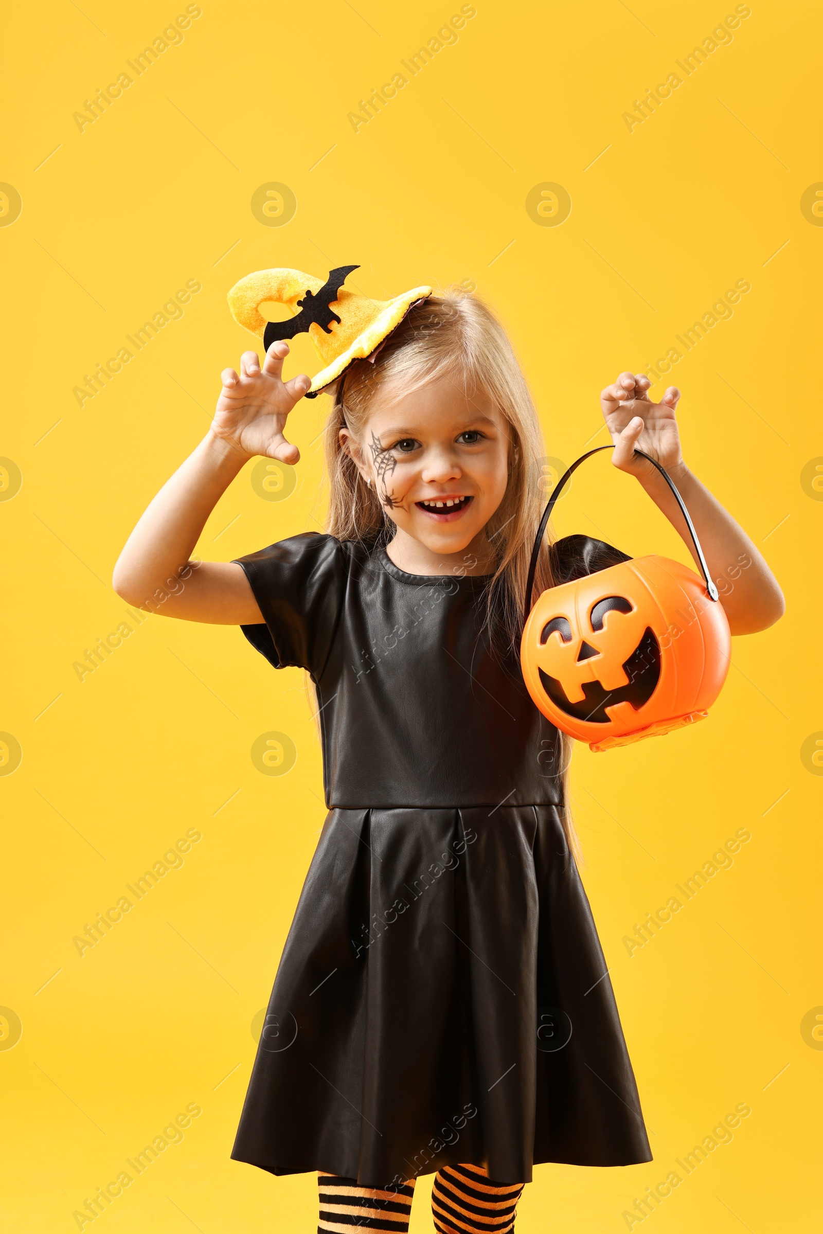 Photo of Cute girl with pumpkin bucket dressed like witch on yellow background. Halloween celebration