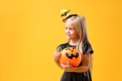 Cute girl with pumpkin bucket dressed like witch on yellow background, space for text. Halloween celebration