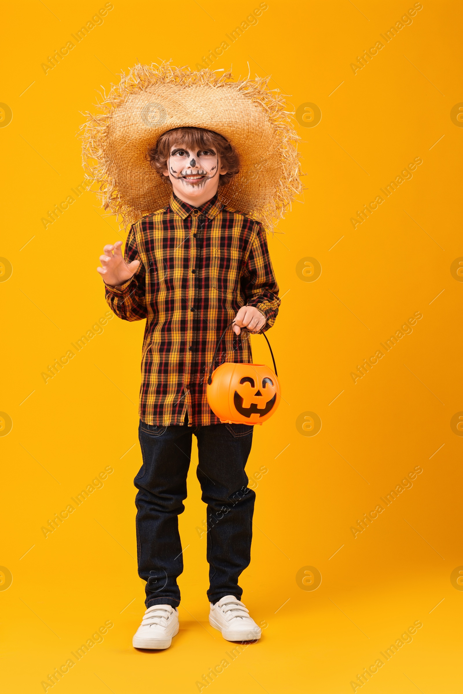 Photo of Funny boy with pumpkin bucket dressed like scarecrow on yellow background. Halloween celebration