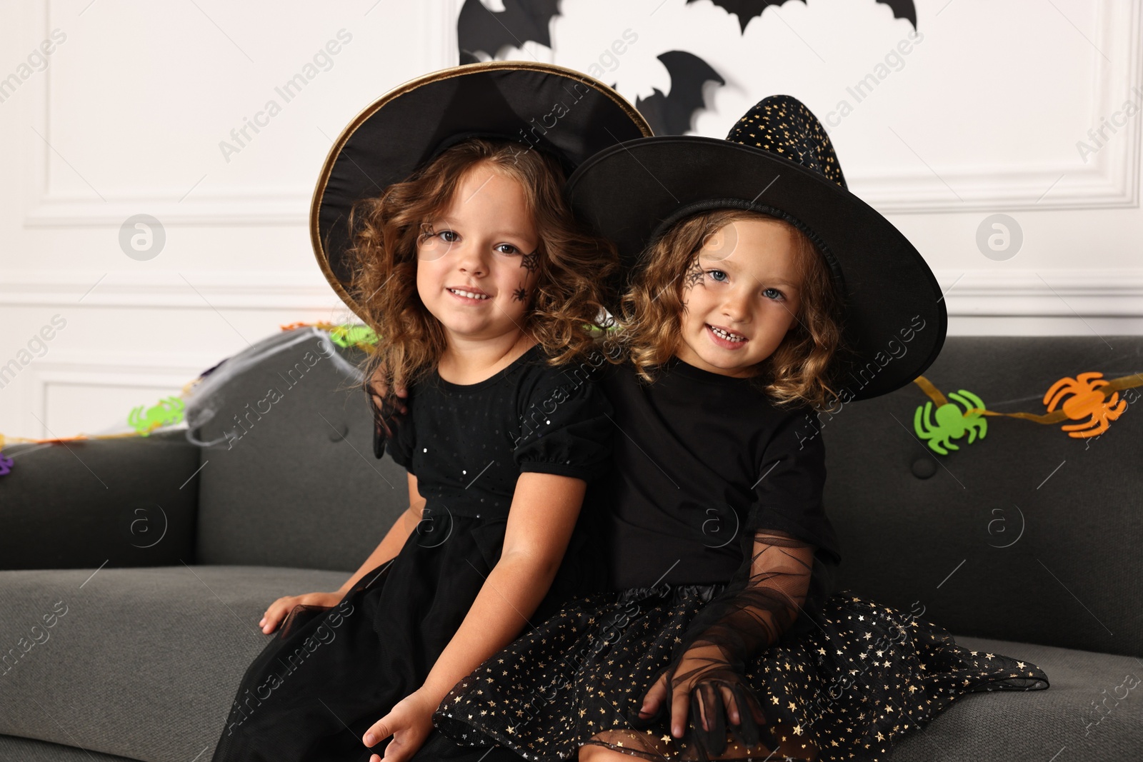 Photo of Cute girls wearing costumes on sofa indoors. Halloween celebration