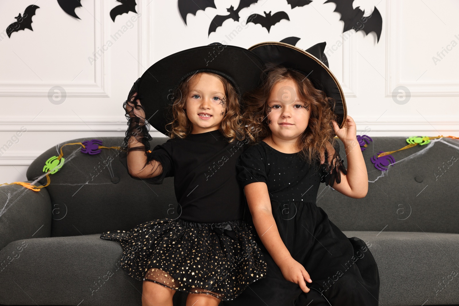 Photo of Cute girls wearing costumes on sofa indoors. Halloween celebration