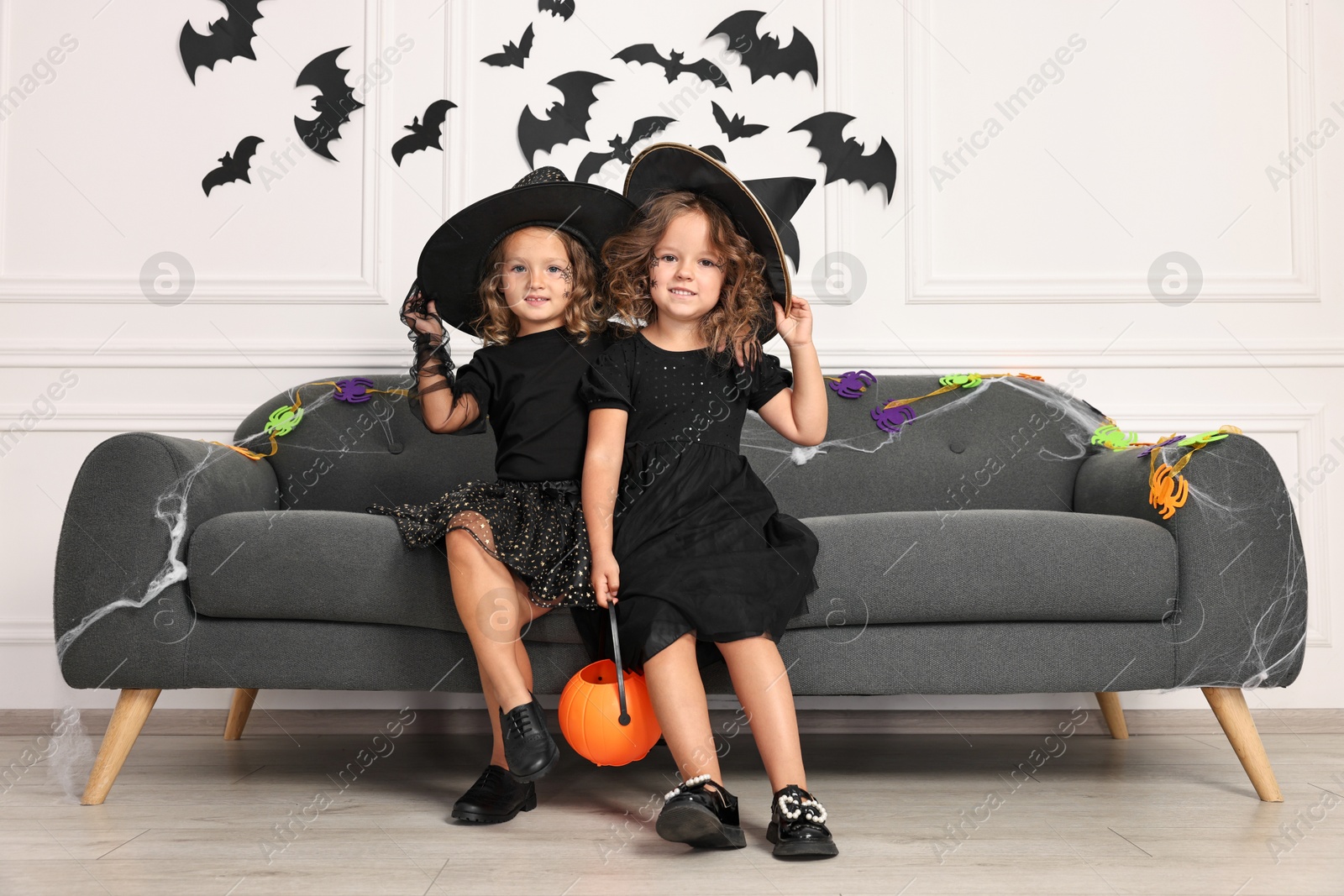 Photo of Cute girls with pumpkin bucket wearing costumes on sofa indoors. Halloween celebration