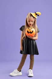 Photo of Cute girl with pumpkin bucket dressed like witch on violet background. Halloween celebration