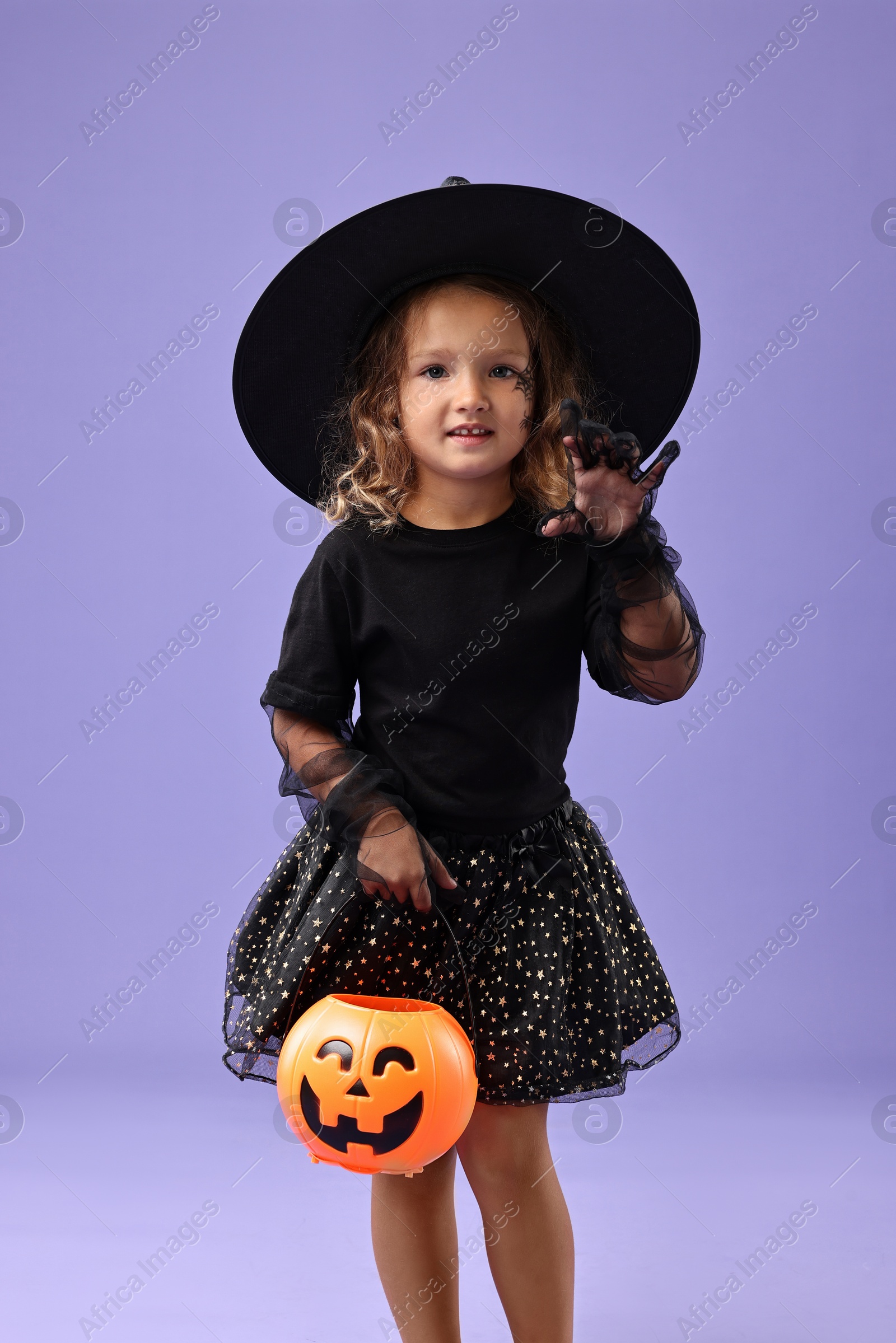 Photo of Cute girl with pumpkin bucket dressed like witch on violet background. Halloween costume