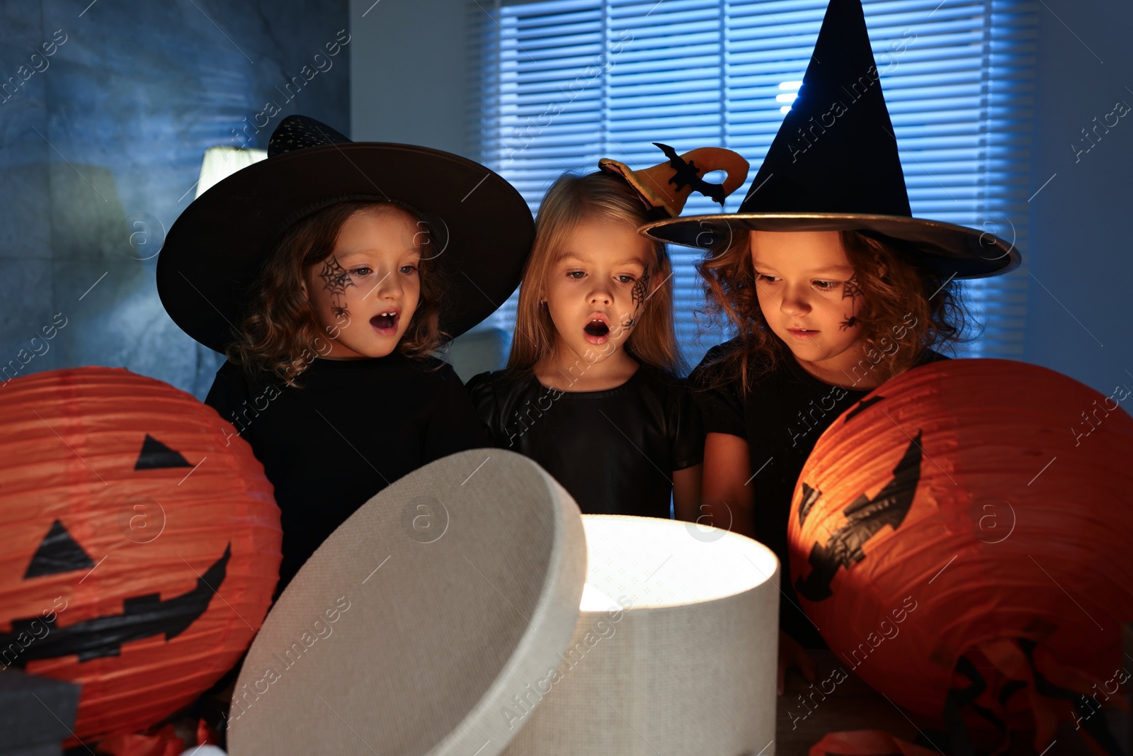 Photo of Surprised children in costumes with festive decor and gift box indoors at night. Halloween celebration