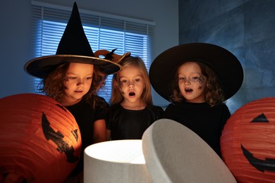 Photo of Surprised children in costumes with festive decor and gift box indoors at night. Halloween celebration