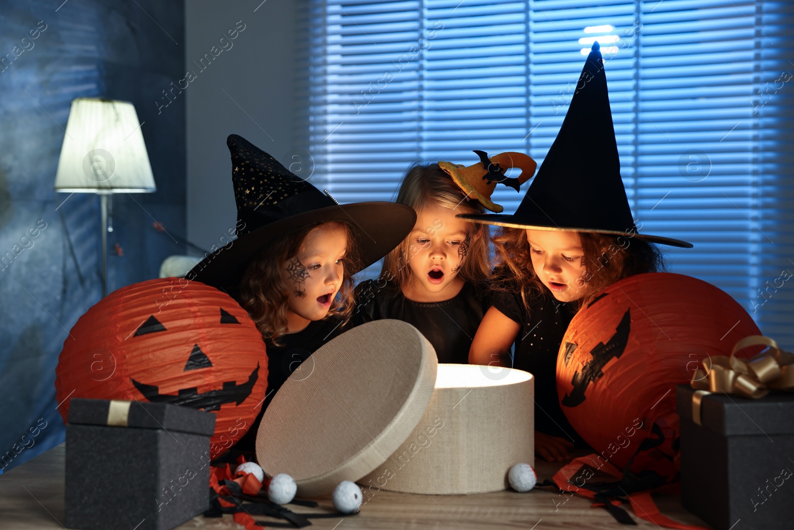 Photo of Surprised children in costumes with festive decor and gift boxes indoors at night. Halloween celebration