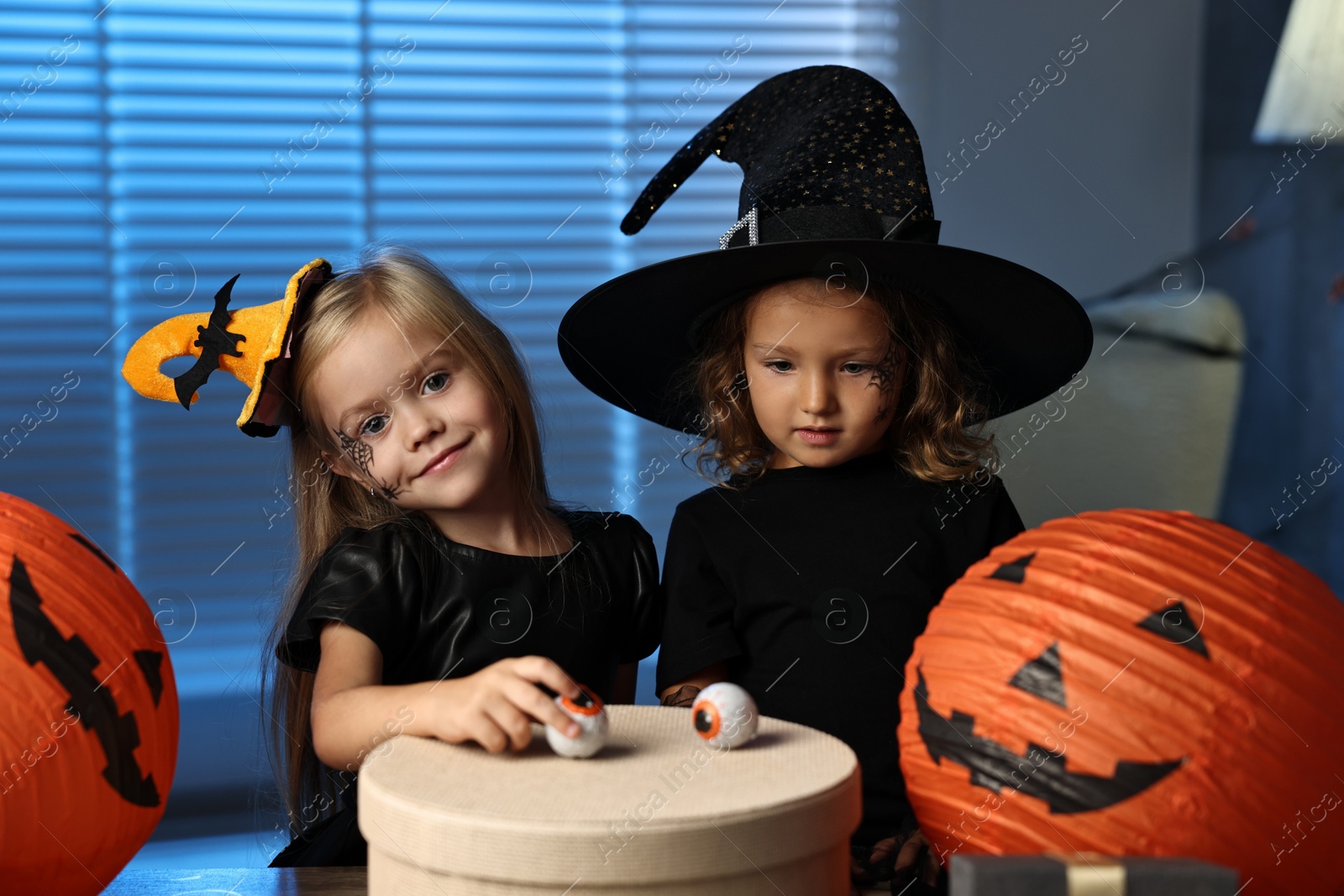 Photo of Cute children in costumes with festive decor and gift box indoors at night. Halloween celebration