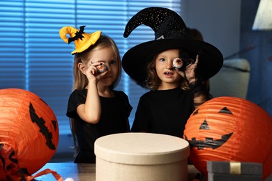 Cute children in costumes with decorative eyeballs, festive decor and gift boxes indoors at night. Halloween celebration