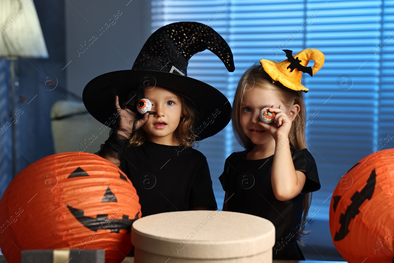 Photo of Cute children in costumes with decorative eyeballs, festive decor and gift boxes indoors at night. Halloween celebration