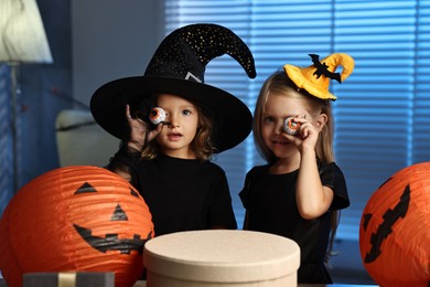 Cute children in costumes with decorative eyeballs, festive decor and gift boxes indoors at night. Halloween celebration