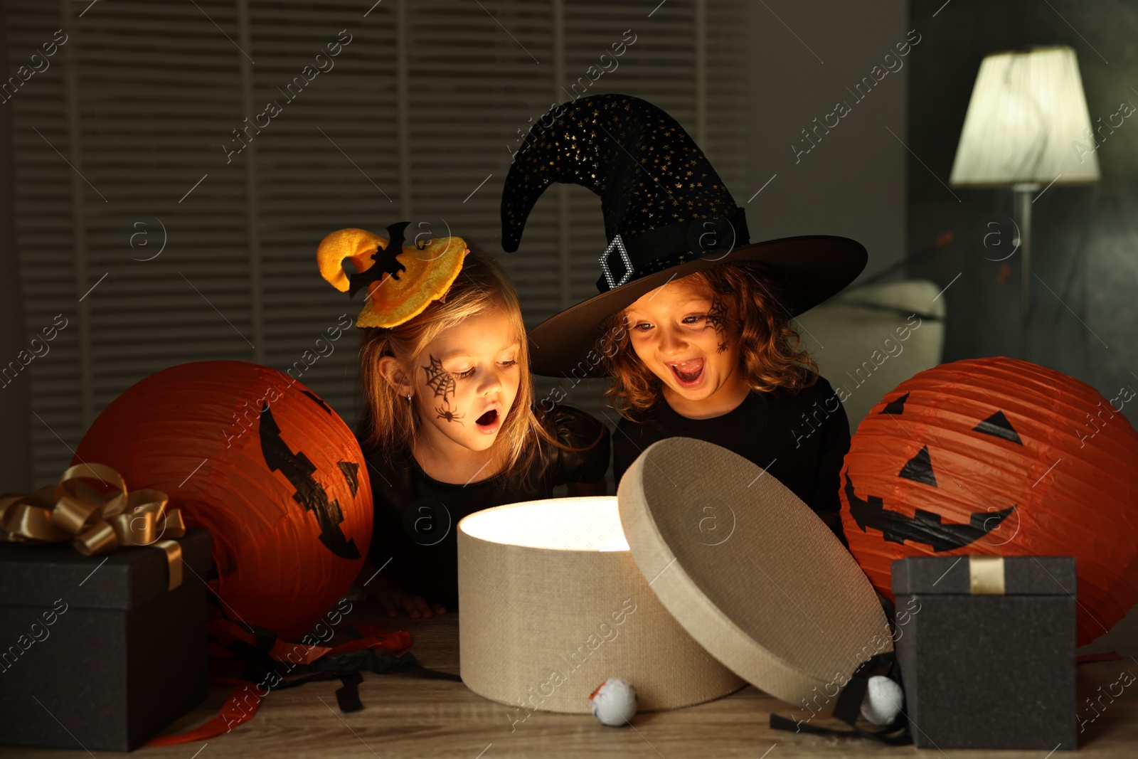 Photo of Surprised children in costumes with festive decor and gift boxes indoors at night. Halloween celebration