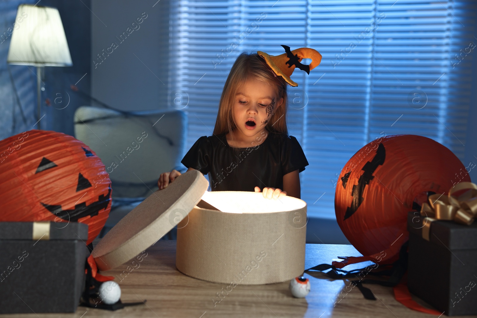 Photo of Surprised girl dressed like witch with festive decor and gift boxes indoors at night. Halloween celebration