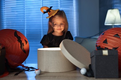 Photo of Cute girl dressed like witch with festive decor and gift boxes indoors at night. Halloween celebration