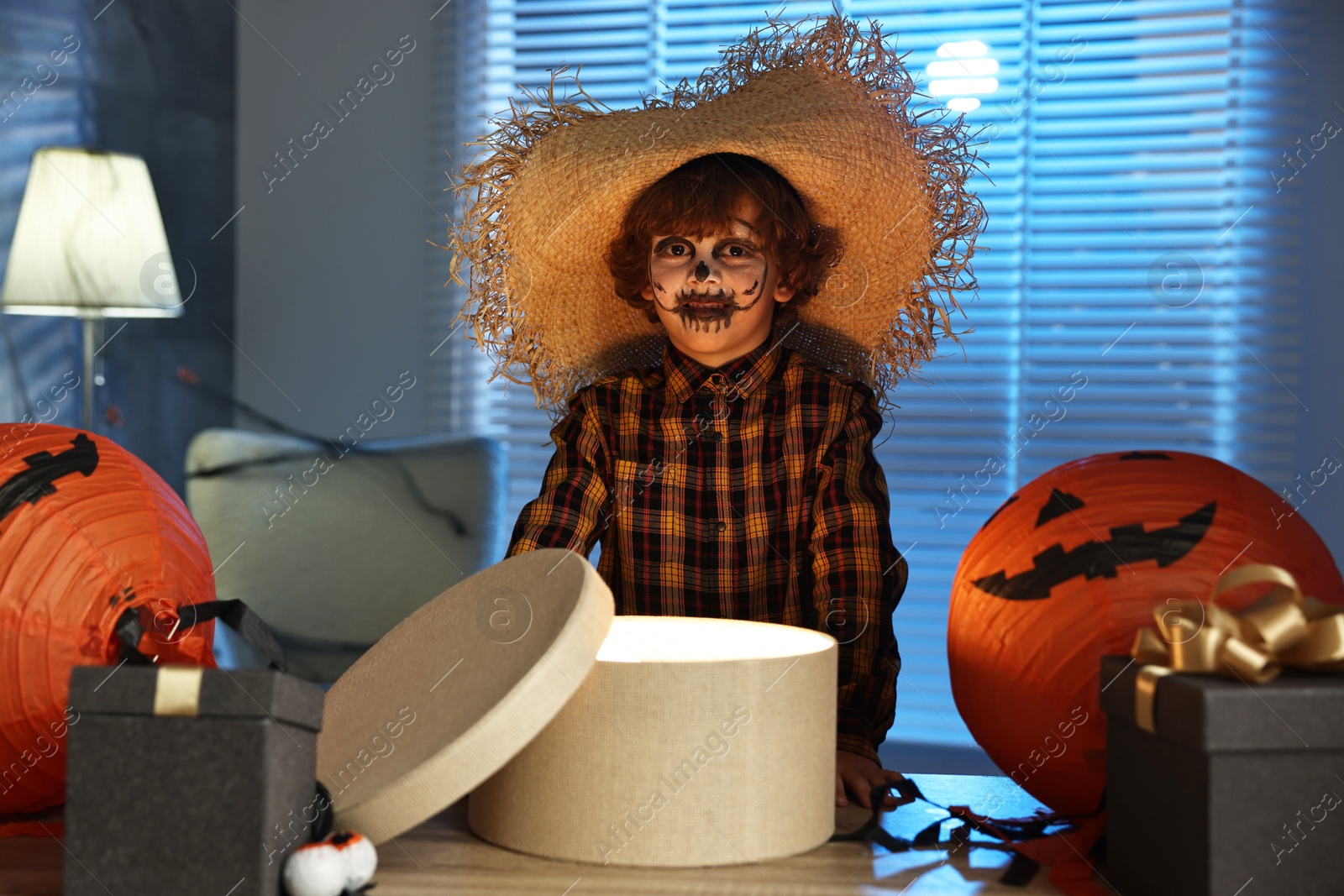 Photo of Cute boy dressed like scarecrow with festive decor and gift boxes indoors at night. Halloween celebration