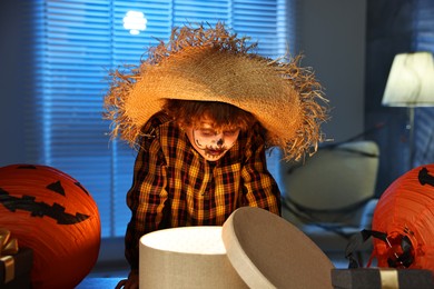 Photo of Cute boy dressed like scarecrow with festive decor and gift boxes indoors at night. Halloween celebration