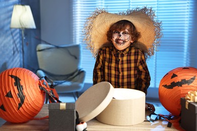 Photo of Cute boy dressed like scarecrow with festive decor and gift boxes indoors at night. Halloween celebration