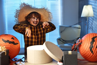 Cute boy dressed like scarecrow with festive decor and gift boxes indoors at night. Halloween celebration