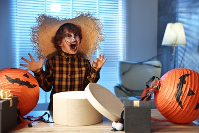 Photo of Cute boy dressed like scarecrow with festive decor and gift boxes indoors at night. Halloween celebration