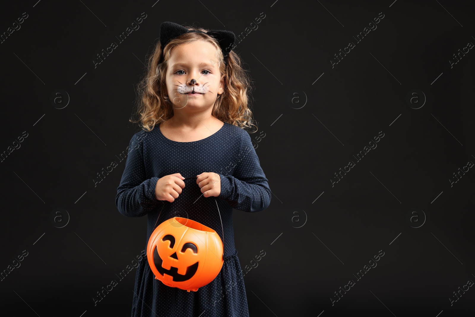 Photo of Cute girl with pumpkin bucket dressed like cat for Halloween celebration on black background, space for text