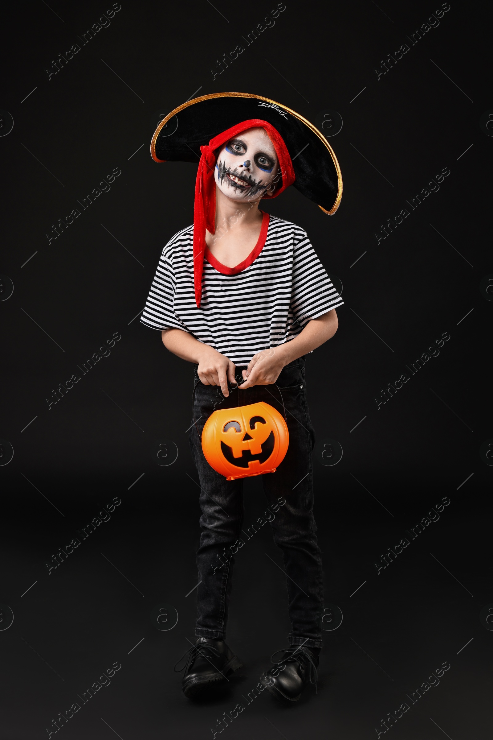 Photo of Funny boy with pumpkin bucket dressed like pirate on black background. Halloween costume