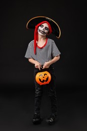 Funny boy with pumpkin bucket dressed like pirate on black background. Halloween costume