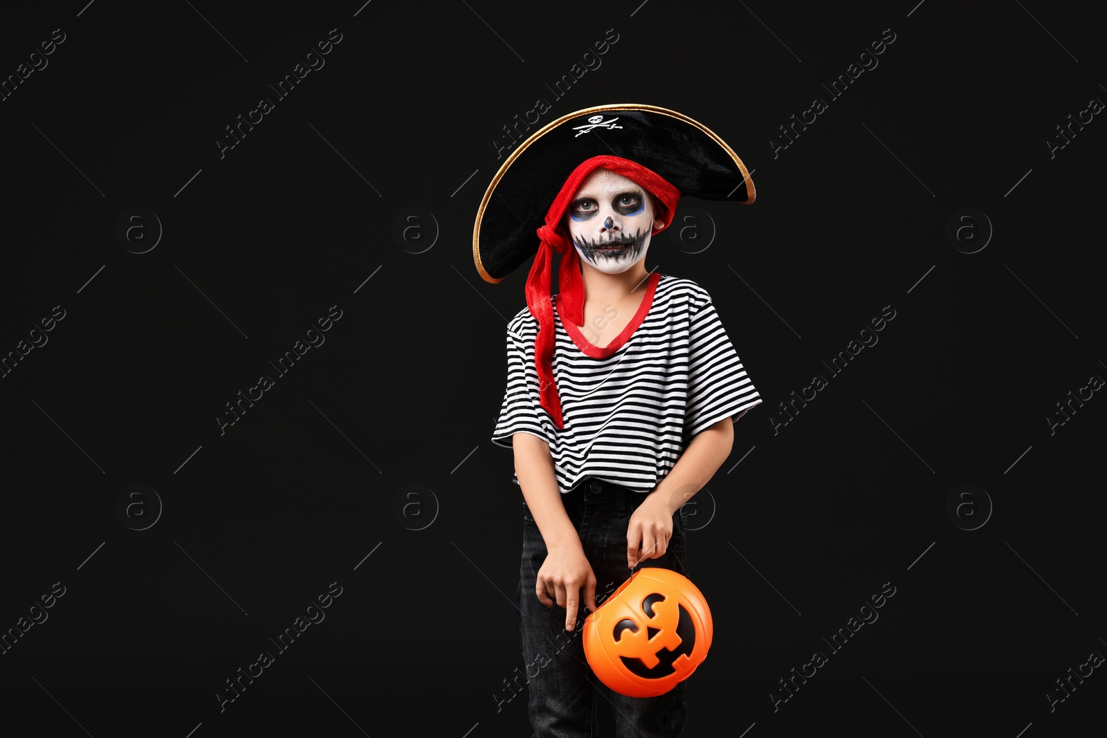 Photo of Funny boy with pumpkin bucket dressed like pirate on black background. Halloween costume