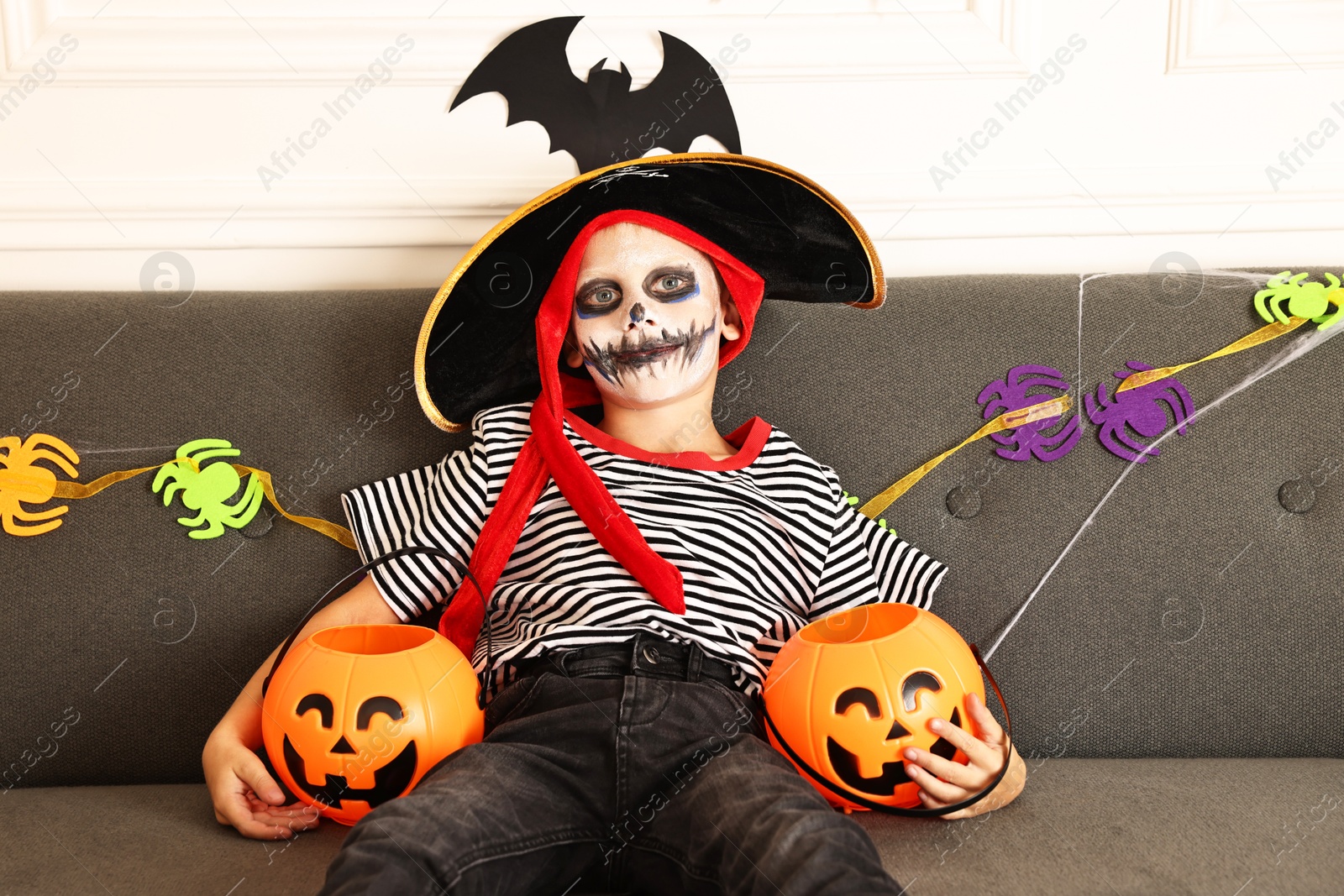 Photo of Funny boy with different Halloween decor dressed like pirate on sofa indoors