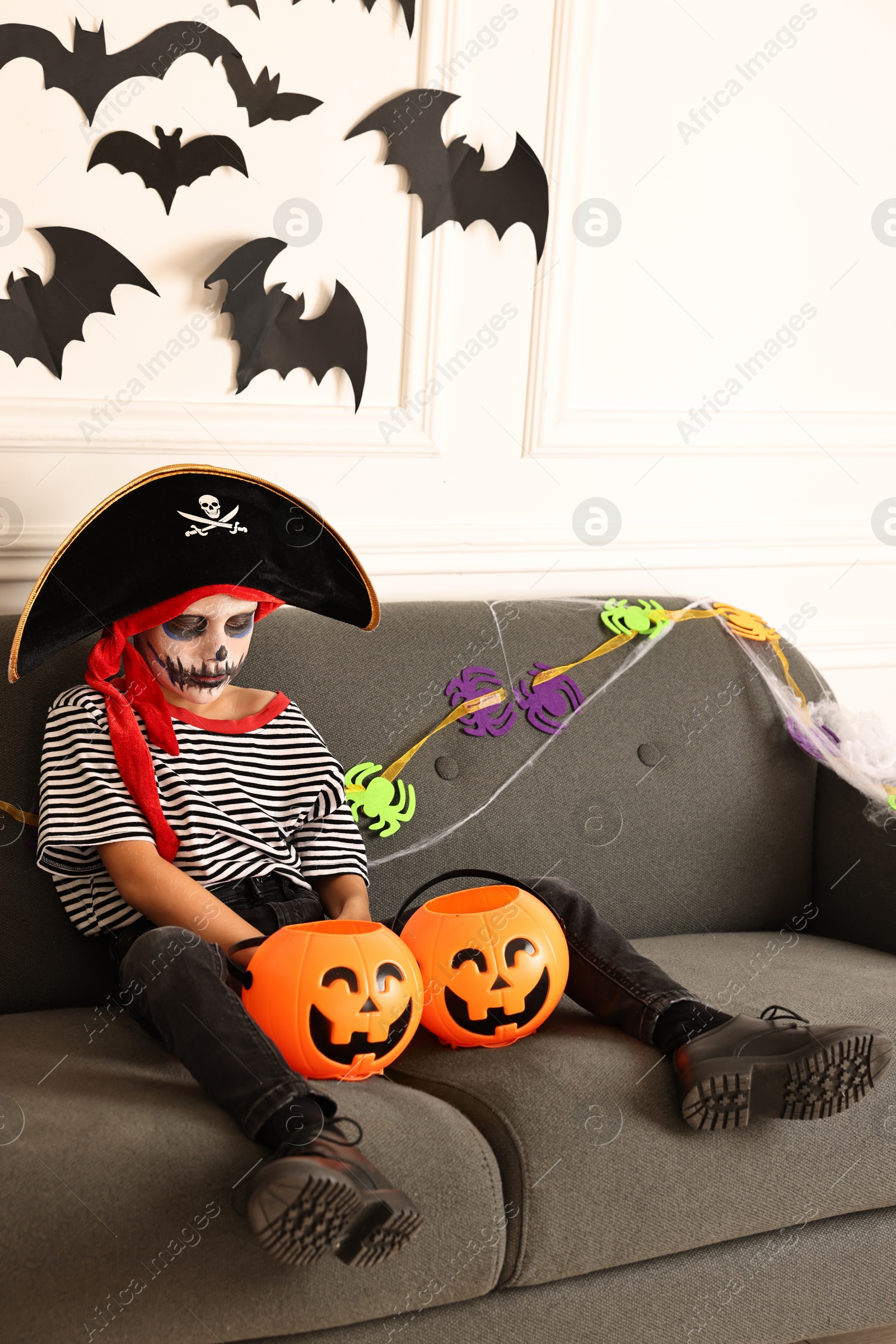 Photo of Funny boy with Halloween decor dressed like pirate on sofa indoors