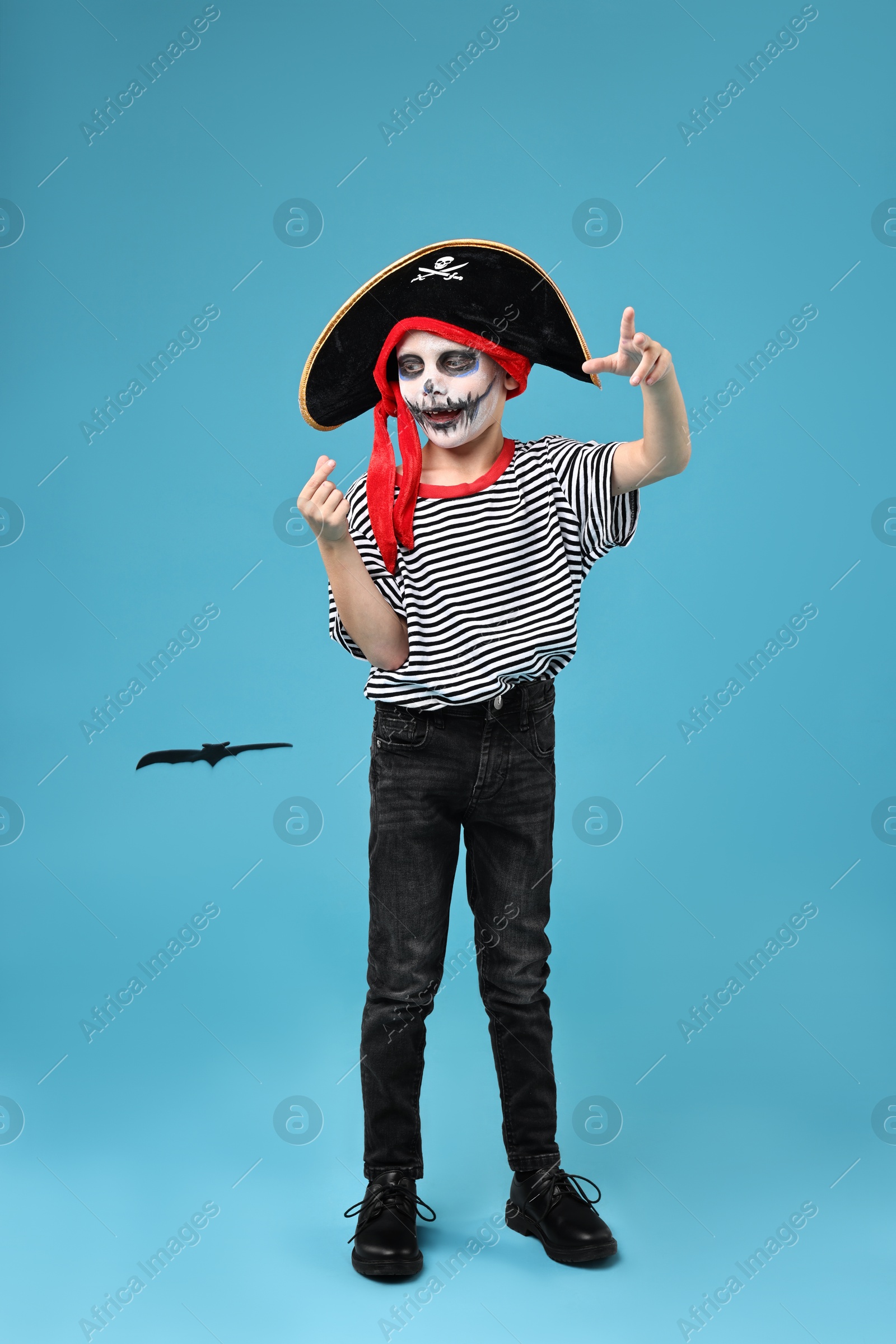 Photo of Funny boy with paper bat dressed like pirate on light blue background. Halloween costume