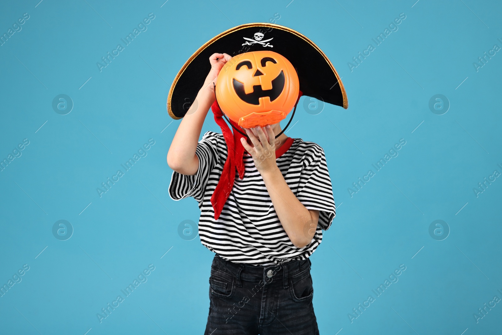 Photo of Boy with pumpkin bucket dressed like pirate on light blue background. Halloween costume