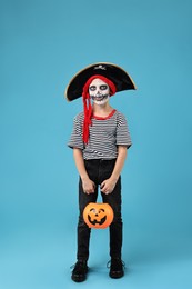 Photo of Funny boy with pumpkin bucket dressed like pirate on light blue background. Halloween costume