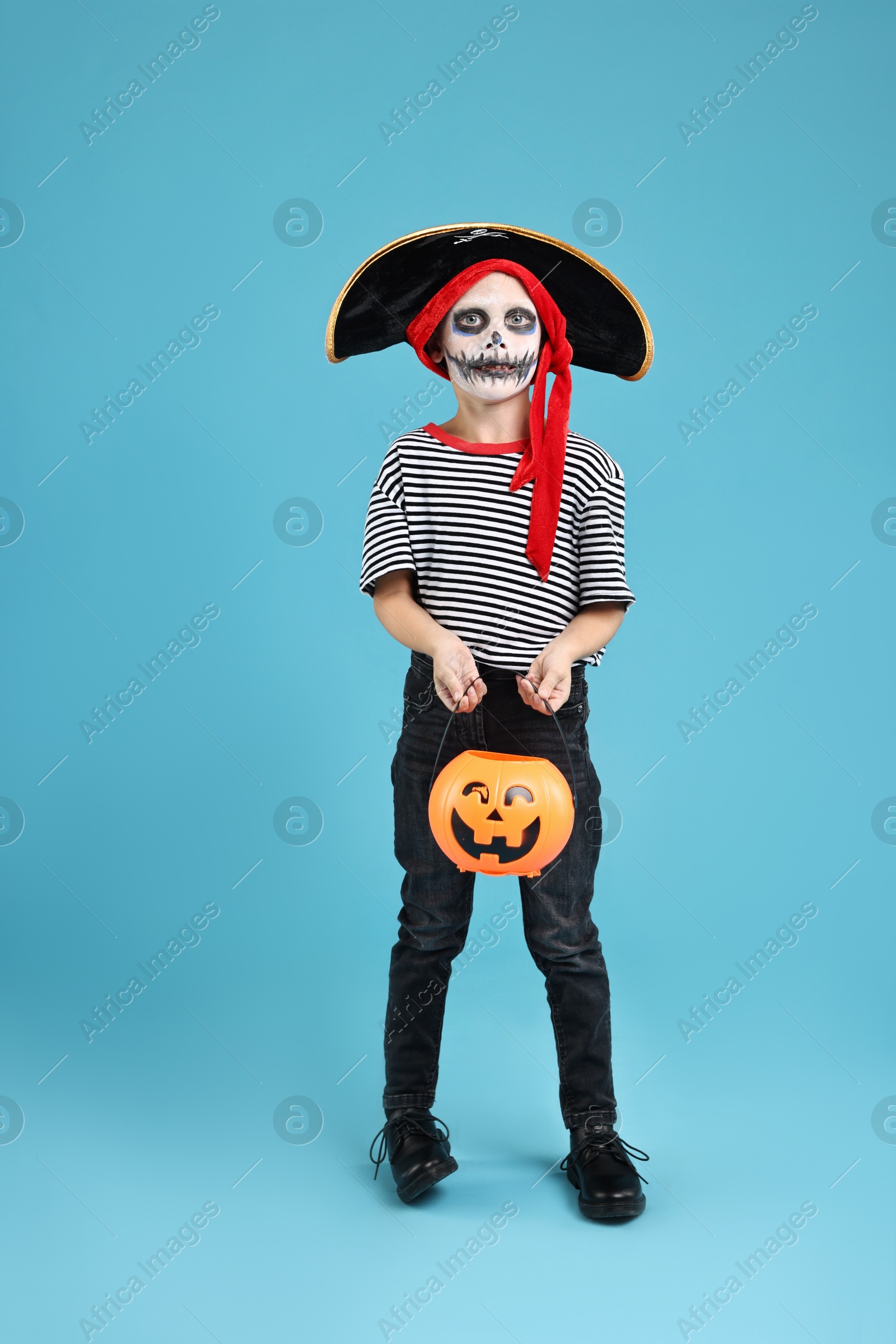 Photo of Funny boy with pumpkin bucket dressed like pirate on light blue background. Halloween costume