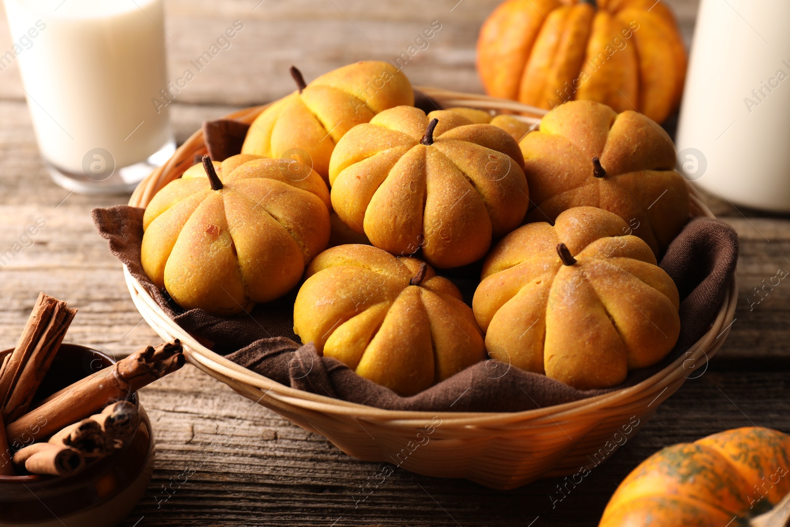 Photo of Wicker basket with tasty pumpkin shaped buns and cinnamon sticks on wooden table, closeup