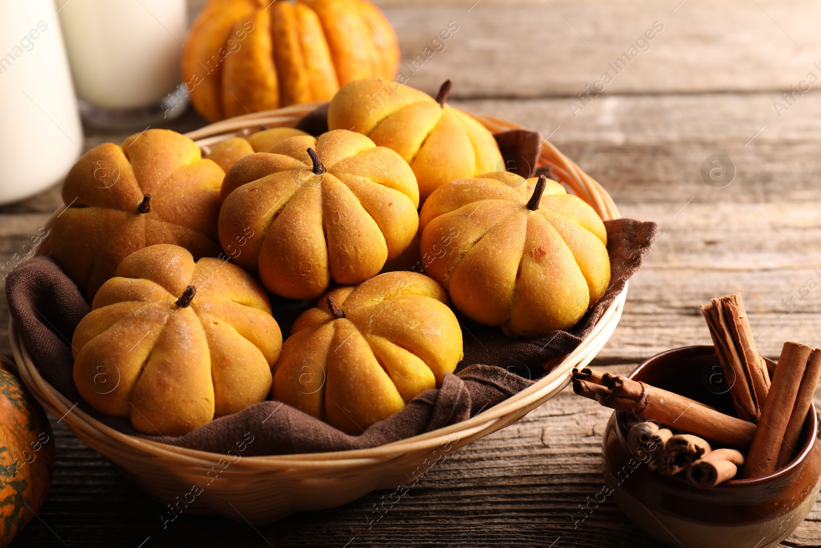 Photo of Wicker basket with tasty pumpkin shaped buns and cinnamon sticks on wooden table, closeup