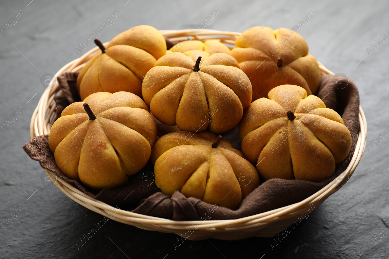 Photo of Wicker basket with tasty pumpkin shaped buns on dark textured table, closeup