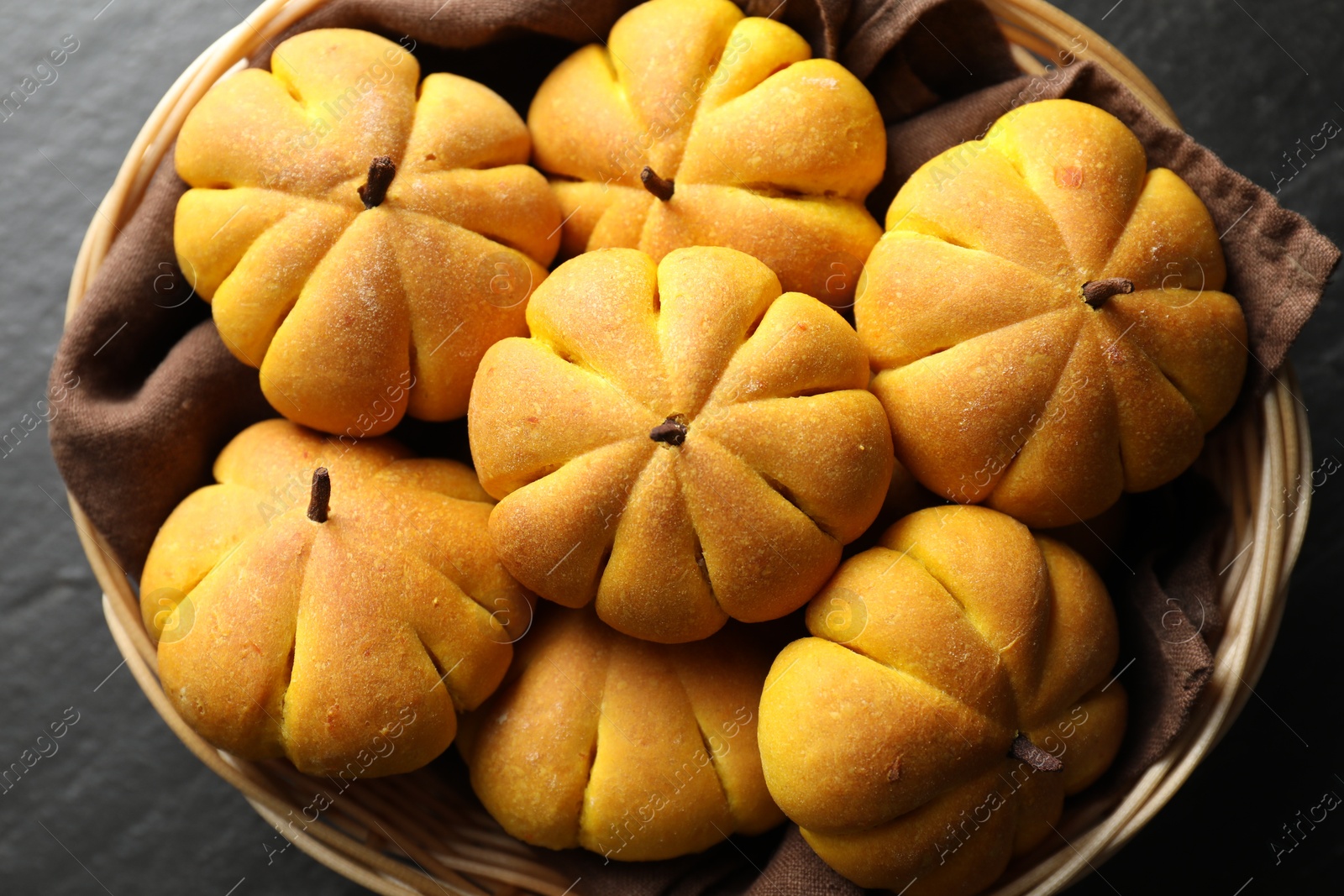 Photo of Wicker basket with tasty pumpkin shaped buns on dark textured table, above view