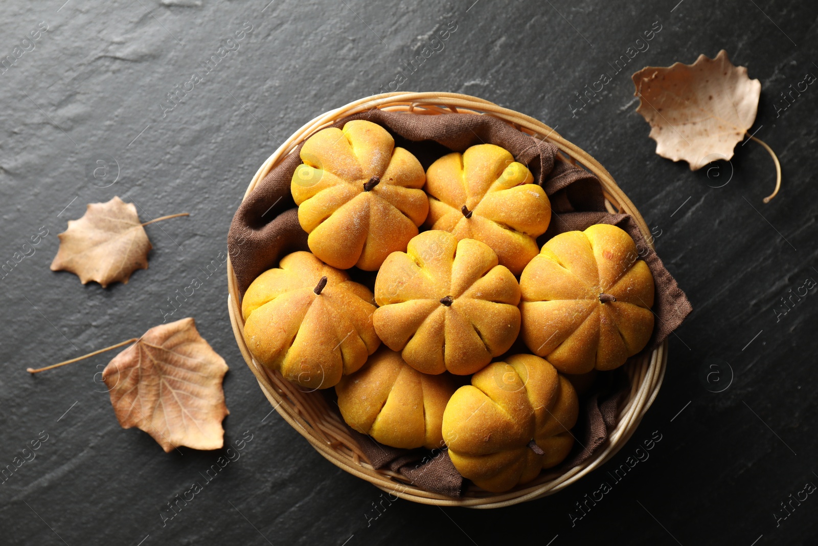 Photo of Wicker basket with tasty pumpkin shaped buns and dry leaves on dark textured table, flat lay