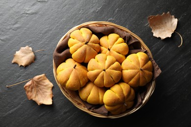 Photo of Wicker basket with tasty pumpkin shaped buns and dry leaves on dark textured table, flat lay