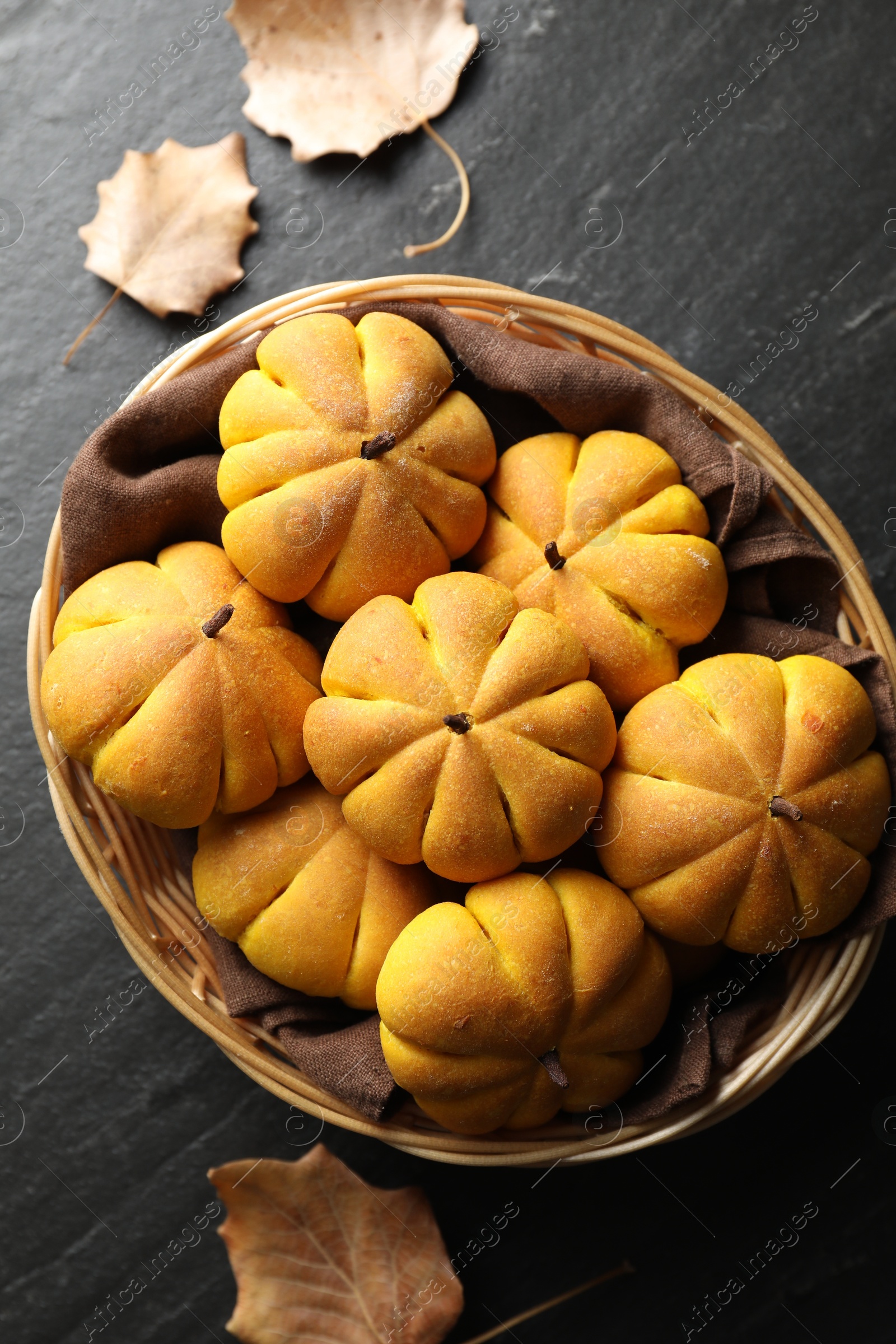 Photo of Wicker basket with tasty pumpkin shaped buns and dry leaves on dark textured table, flat lay