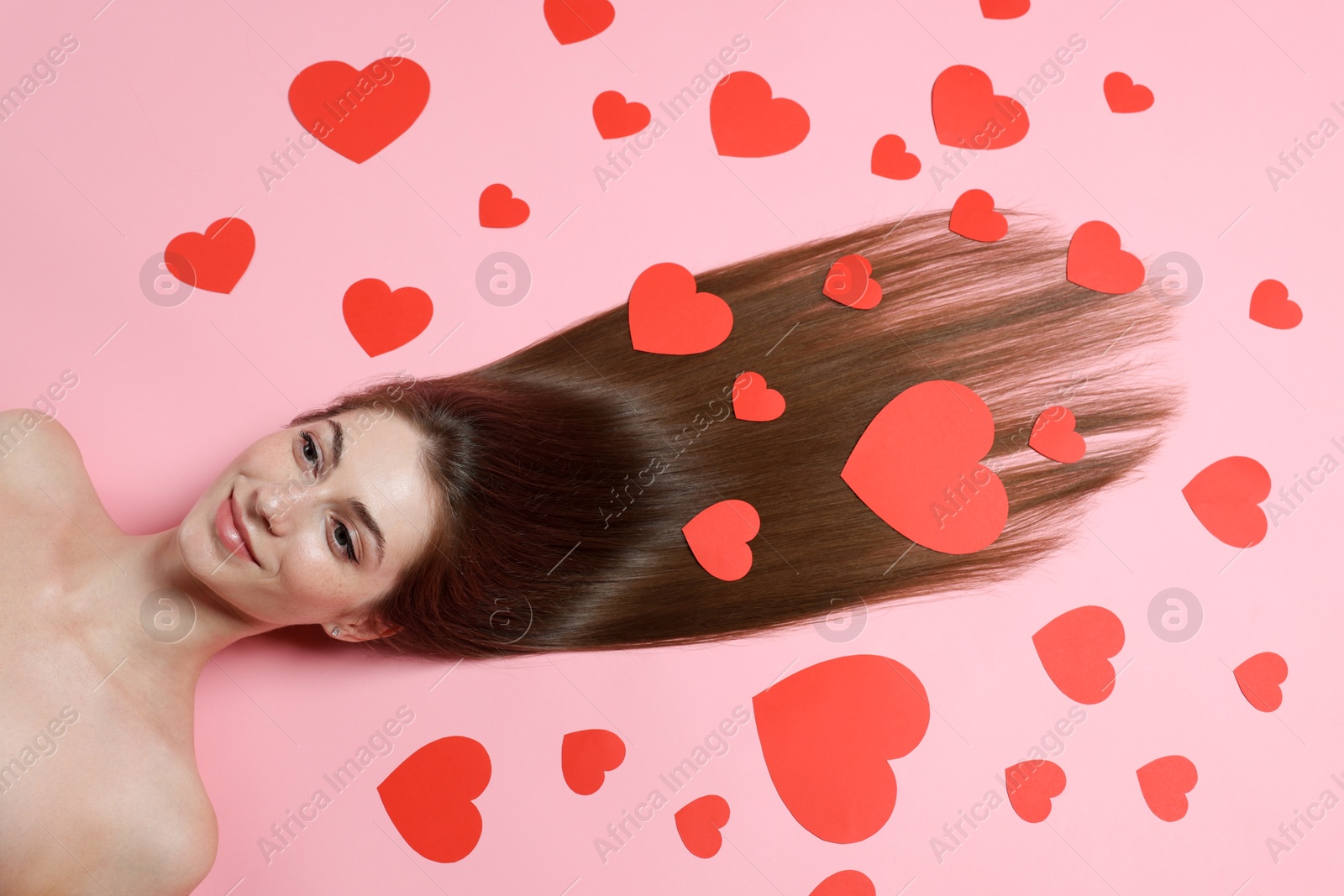 Photo of Beautiful young woman with long hair and red paper hearts on pink background, top view