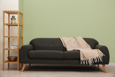 Stylish sofa with blanket and shelving unit near green wall indoors