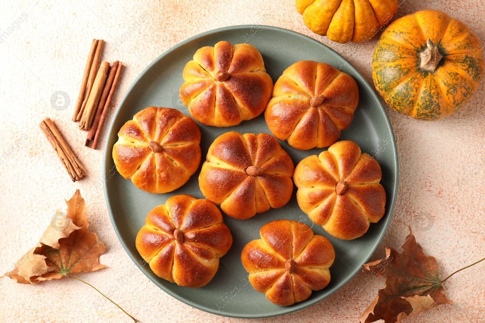 Photo of Tasty pumpkin shaped buns, ingredients and dry leaves on beige textured table, flat lay