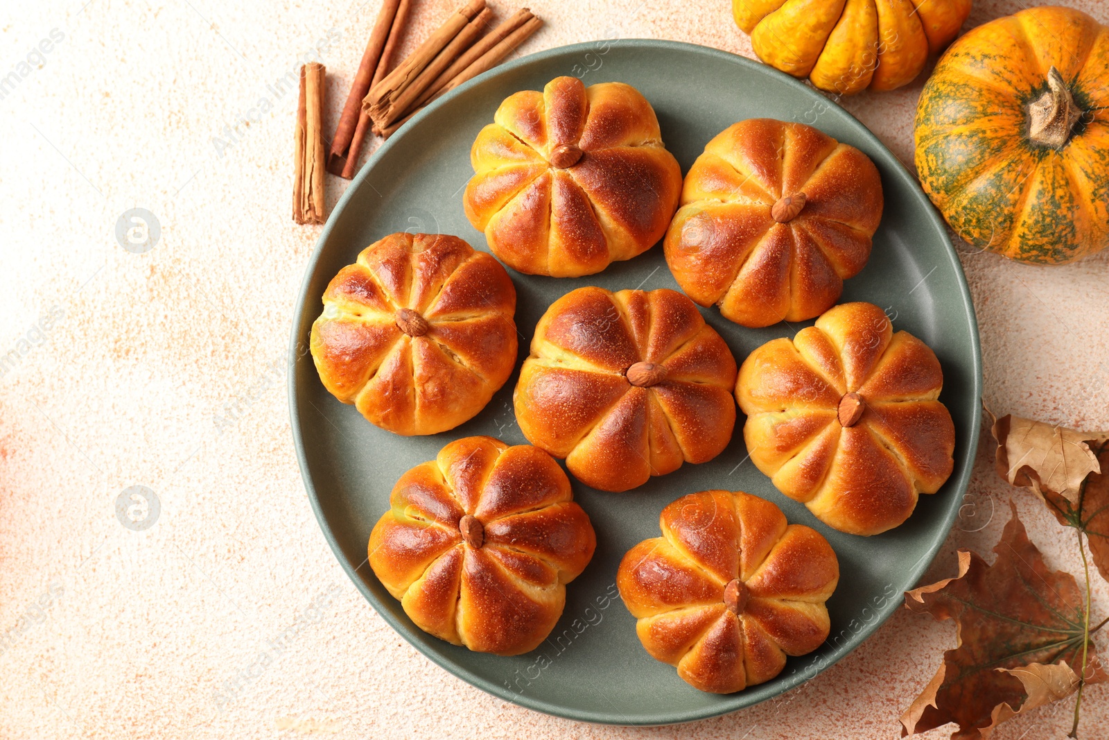 Photo of Tasty pumpkin shaped buns, ingredients and dry leaves on beige textured table, flat lay