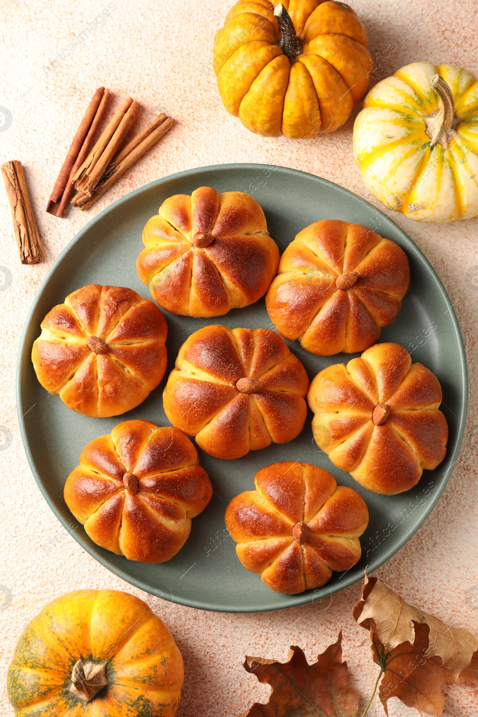 Photo of Tasty pumpkin shaped buns, ingredients and dry leaves on beige textured table, flat lay